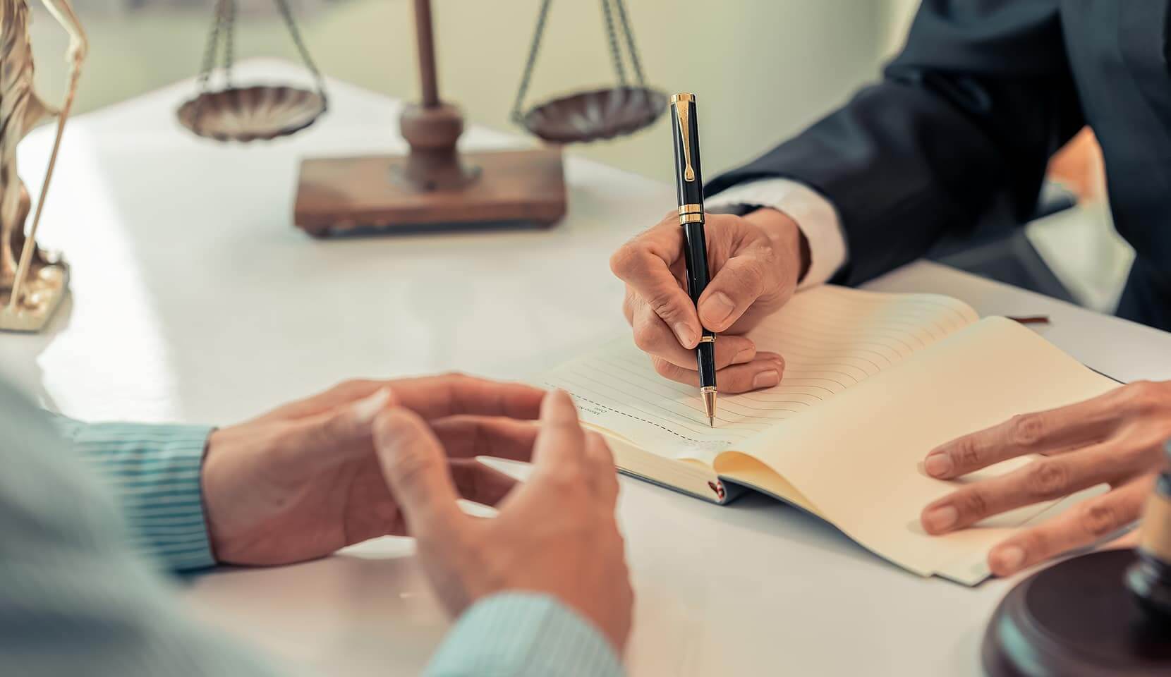 Lawyer writing in a notebook during a legal consultation with a client, with scales of justice and a judge's gavel on the desk
